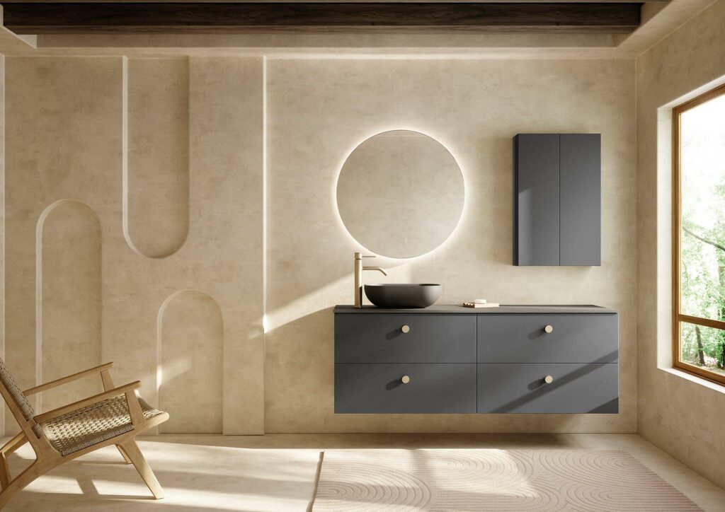 Mueble de baño suspendido con dos líneas de cajones dobles en color gris mate, acompañado de una columna de almacenaje a juego, un espejo redondo con luz incluída, lavabo sobre encimera negro mate redondo y grifo sobre encimera en color oro cepillado, colocado todo sobre pared de cerámica en color tierra natural